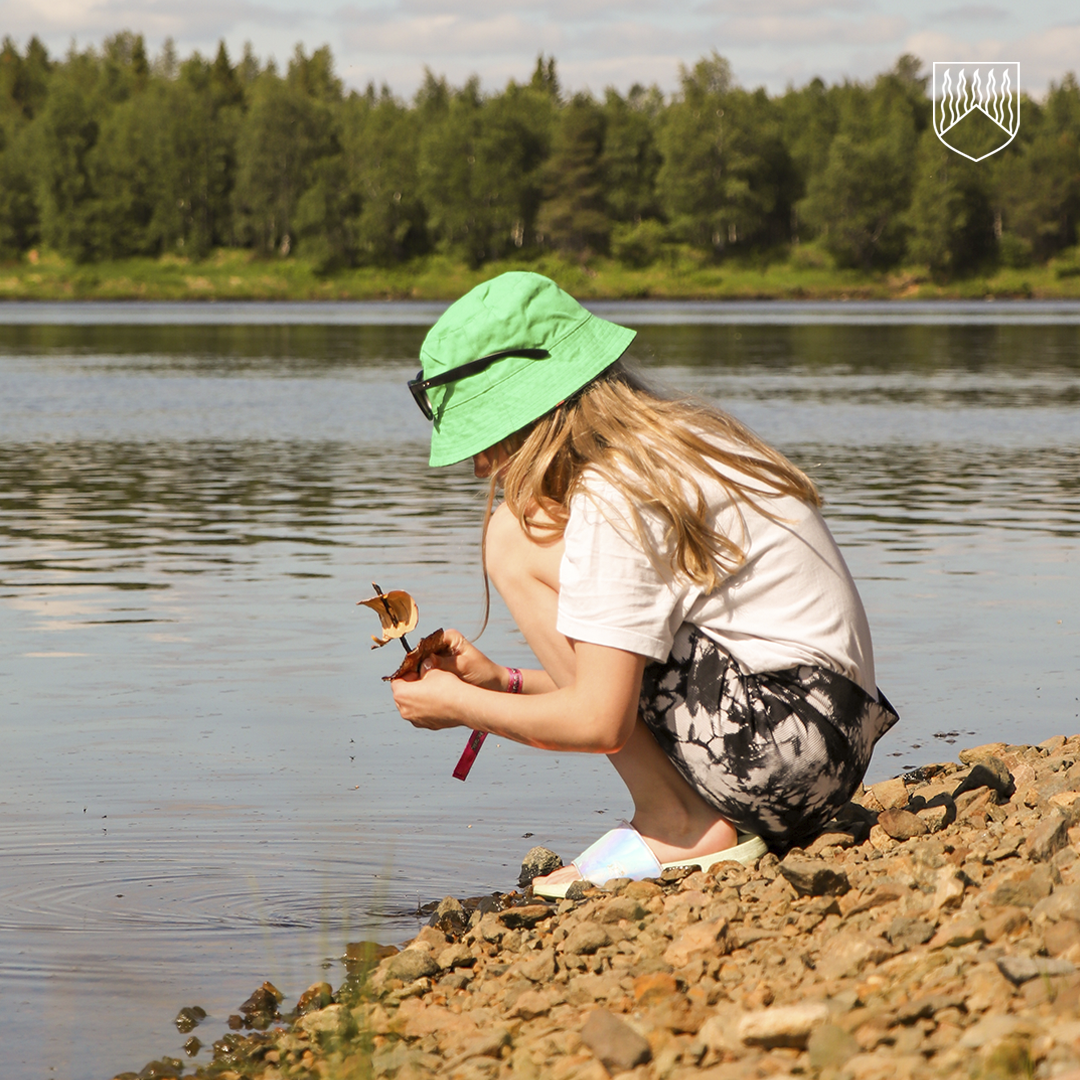 Kuvassa tyttö joen rannalla laskemassa veteen kaarnavenettä. Kuvan ottanut Markku Hamari.