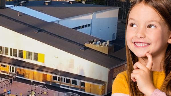 Kuvassa on suuri, moderni koulurakennus, jonka edessä on leikkikenttä ja urheilukenttä. Rakennuksen ympärillä on metsää ja taustalla näkyy laaja metsämaisema. Kuvan etualalla on lapsi, joka hymyilee ja katsoo ylöspäin mietteliäänä.