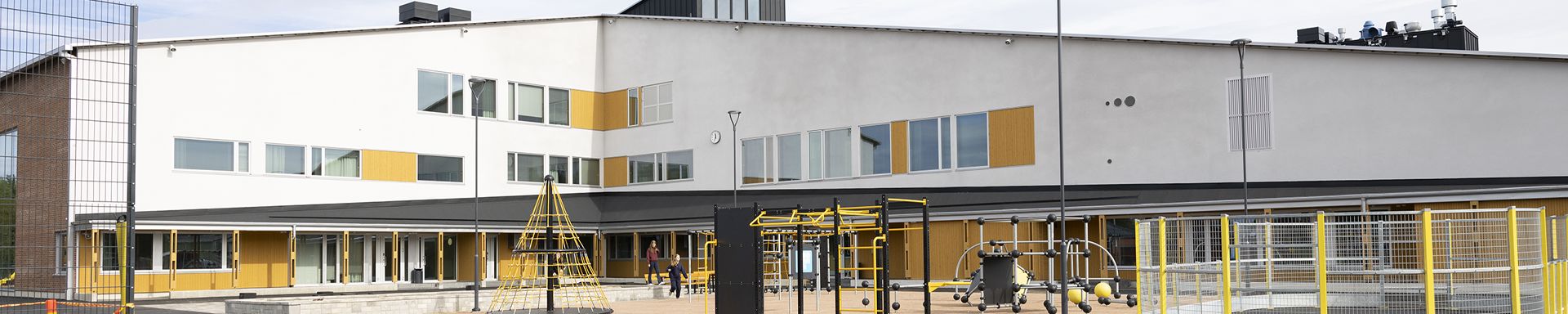 Moderni rakennus, jonka edustalla on leikkipaikka. Rakennuksessa on valkoinen julkisivu ja keltaisia yksityiskohtia ikkunoiden ja ovien ympärillä. Leikkivälineisiin kuuluu kiipeilytelineitä, keinuja ja muita laitteita, joissa on keltaisia osia. Alue on aidattu keltaisin tolpin, ja kokonaisuus viittaa koulun tai oppilaitoksen ympäristöön.