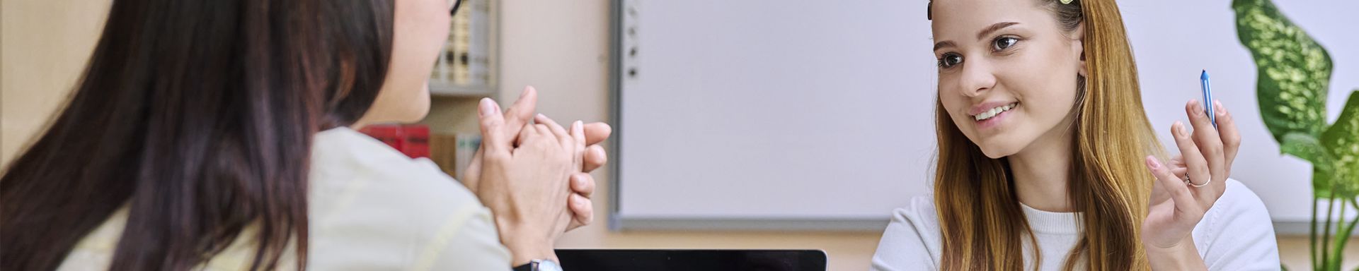 Kaksi henkilöä istuu vastakkain pöydän ääressä. Toinen pitelee kynää ja elehtii kädellään, kun taas toinen pitää käsiään yhdessä. Taustalla näkyy valkotaulu ja kirjahyllyjä. Ympäristö vaikuttaa toimisto- tai luokkahuoneelta.