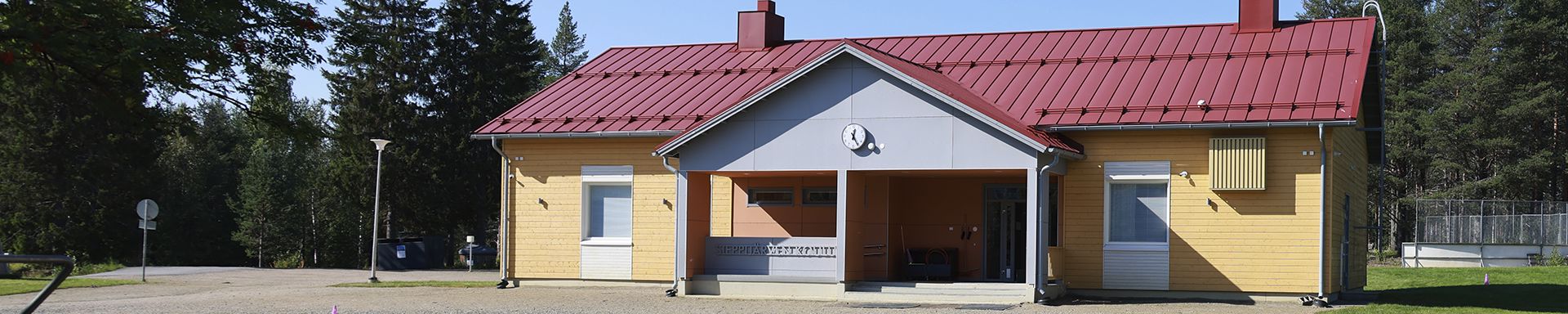 Yksikerroksinen rakennus, jossa on punainen metallikatto ja vaaleankeltaiset puuverhoillut seinät. Rakennuksessa on valkoiset ikkunankarmit ja keskellä sisäänkäynti, jonka yläpuolella on kello. Taustalla näkyy puita, ja sää on kirkas sinitaivainen.