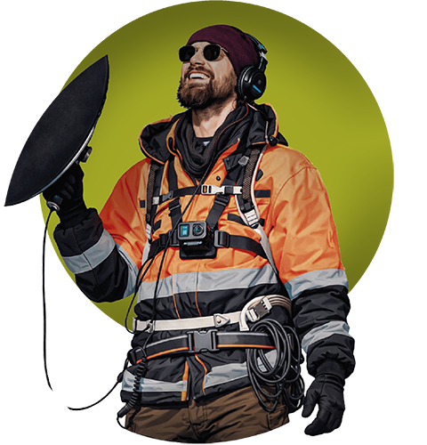 Henkilö seisoo ulkona yllään kirkkaan oranssi heijastava suojatakki ja varustevaljaat. Hänellä on päässään kuulokkeet ja rinnassaan pieni kamera. Hän pitää kädessään suurta, pyöreää laitetta, joka muistuttaa suuntaavaa mikrofonia tai antennia, ja siitä lähtee johto hänen varusteisiinsa. Henkilö näyttää työskentelevän kenttäolosuhteissa tai teknisessä mittaustehtävässä.