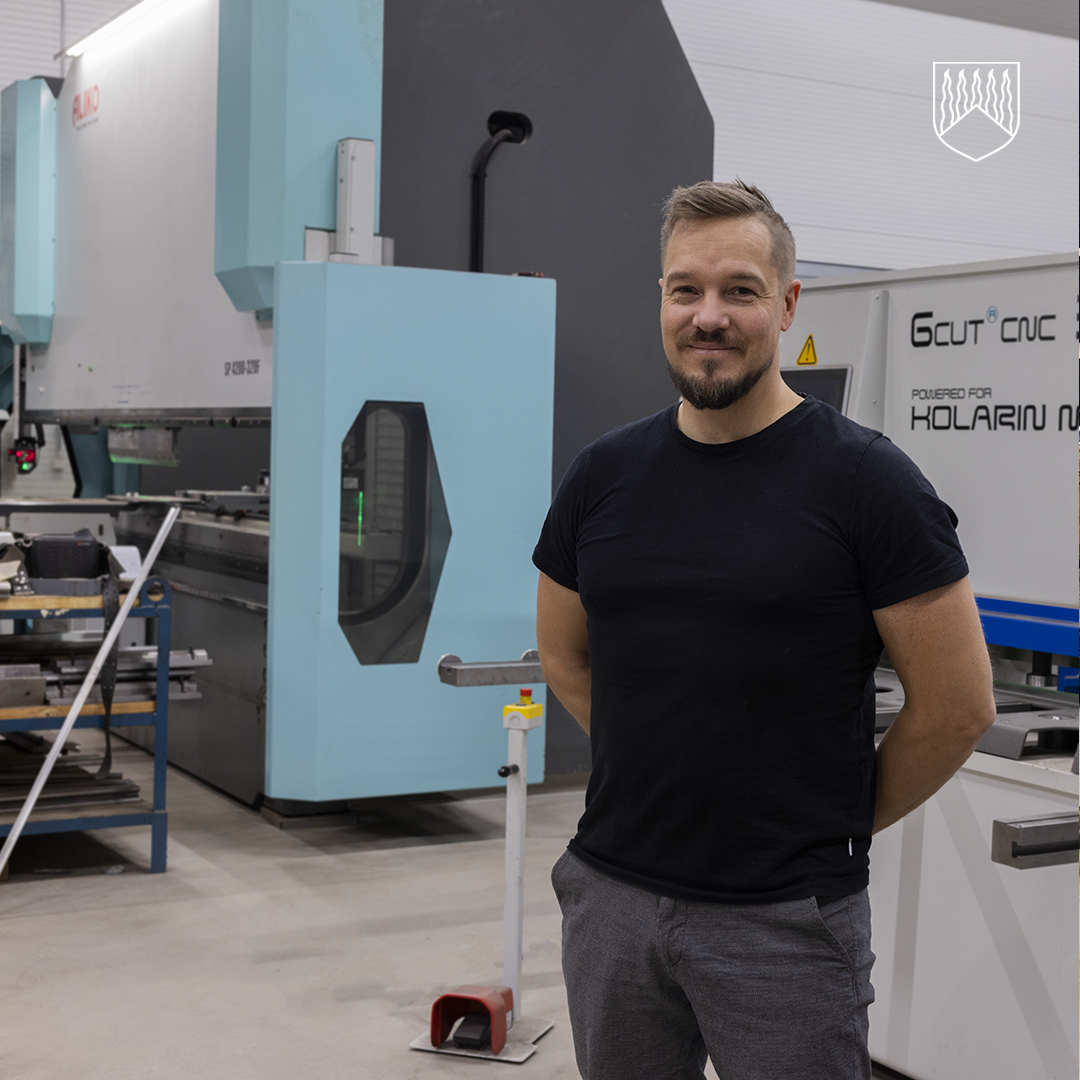 Ari Hooli seisoo metallityöpajassa suuren vaaleansinisen ja harmaan levytyökoneen edessä. Taustalla näkyy muita metallintyöstölaitteita ja työpöytiä. Henkilö on pukeutunut mustaan t-paitaan ja harmaisiin housuihin, kädet selän takana.