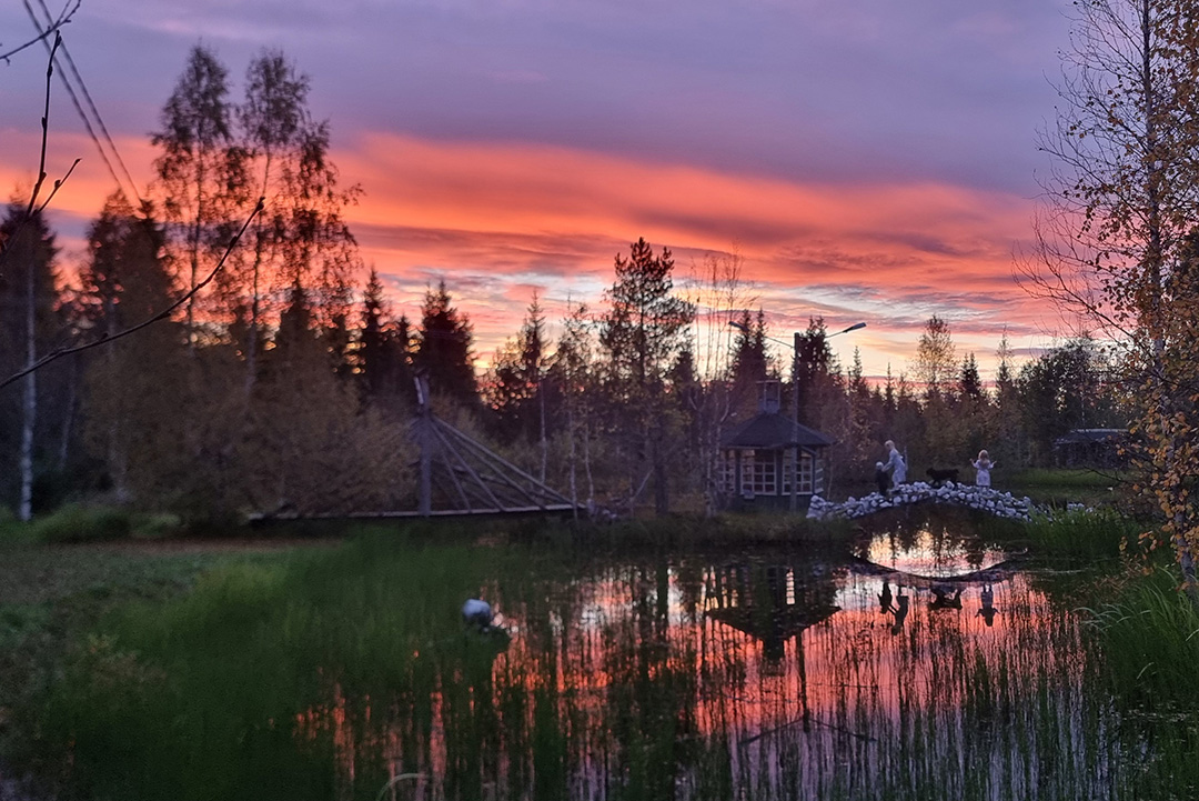 Kuvassa syksyisen maagisissa väreissä lapset sekä koiran tallustelevat puron yli siltaa pitkin.