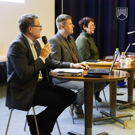 Kolme henkilöä istuu vierekkäin kahden pyöreän pöydän ääressä kokoustilassa. Vasemmalla kunnanjohtaja Markku Vehkaoja pitää mikrofonia ja näyttää olevan puheenvuorossa. Keskellä kunnanvaltuuston puheenjohtaja V-P Uusitalo istuu kannettavan tietokoneen edessä, ja oikealla hallintosihteeri Ulla Liikamaa istuu hieman sivuttain. Pöydillä on kannettavia tietokoneita, muistivihkoja, vesipulloja ja muita pieniä esineitä. Taustalla on tummat verhot ja valkokangas.