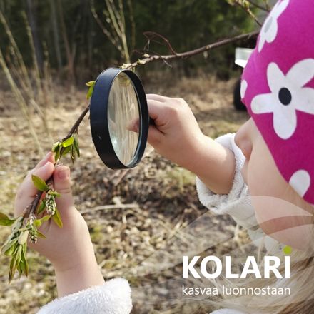 Lapsi tutkii oksaa suurennuslasilla ulkona keväisessä maastossa. Oksassa on vihreitä silmuja, ja taustalla näkyy ruskeaa kuivaa heinikkoa. Lapsella on päässään pinkki pipo, jossa on valkoisia kukkakuvioita.