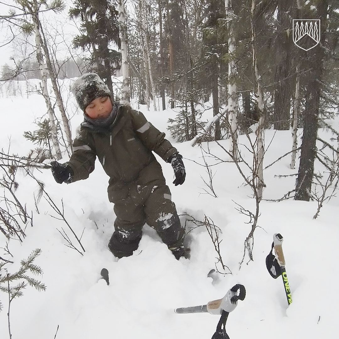 Henkilö seisoo syvässä lumessa metsäisessä maastossa, pukeutuneena paksuun talviasuun ja hansikkaisiin. Etualalla näkyy kaksi hiihtosauvaa, joissa on puiset varret ja metalliset terät, osittain lumen peitossa. Taustalla on lumisia puita ja talvinen maisema.