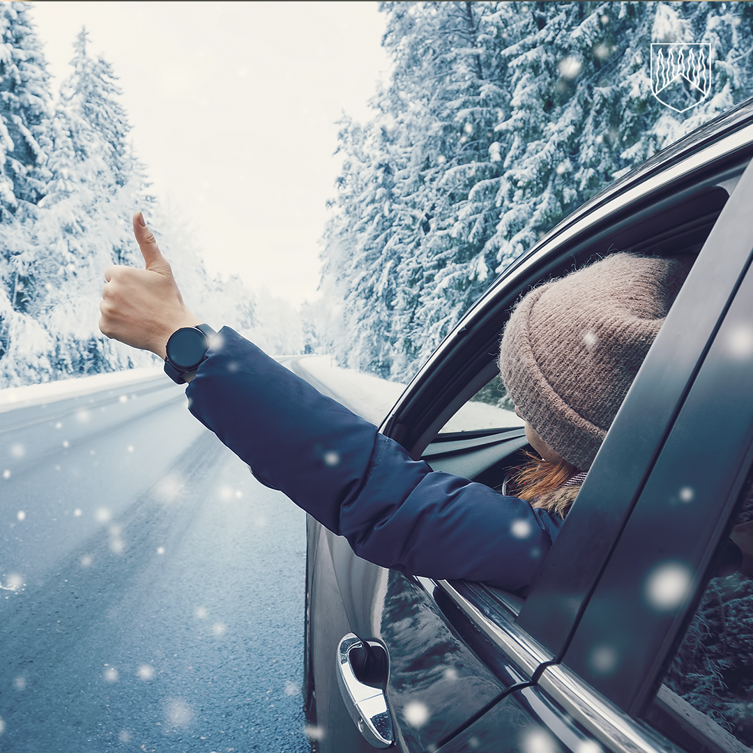 Auto ajaa talvisella, lumisella tiellä, jonka molemmin puolin on lumipeitteisiä kuusia. Auton sivuikkunasta näkyy henkilö, joka ojentaa kätensä ulos ja näyttää peukaloa ylöspäin. Taustalla leijailee lumihiutaleita.