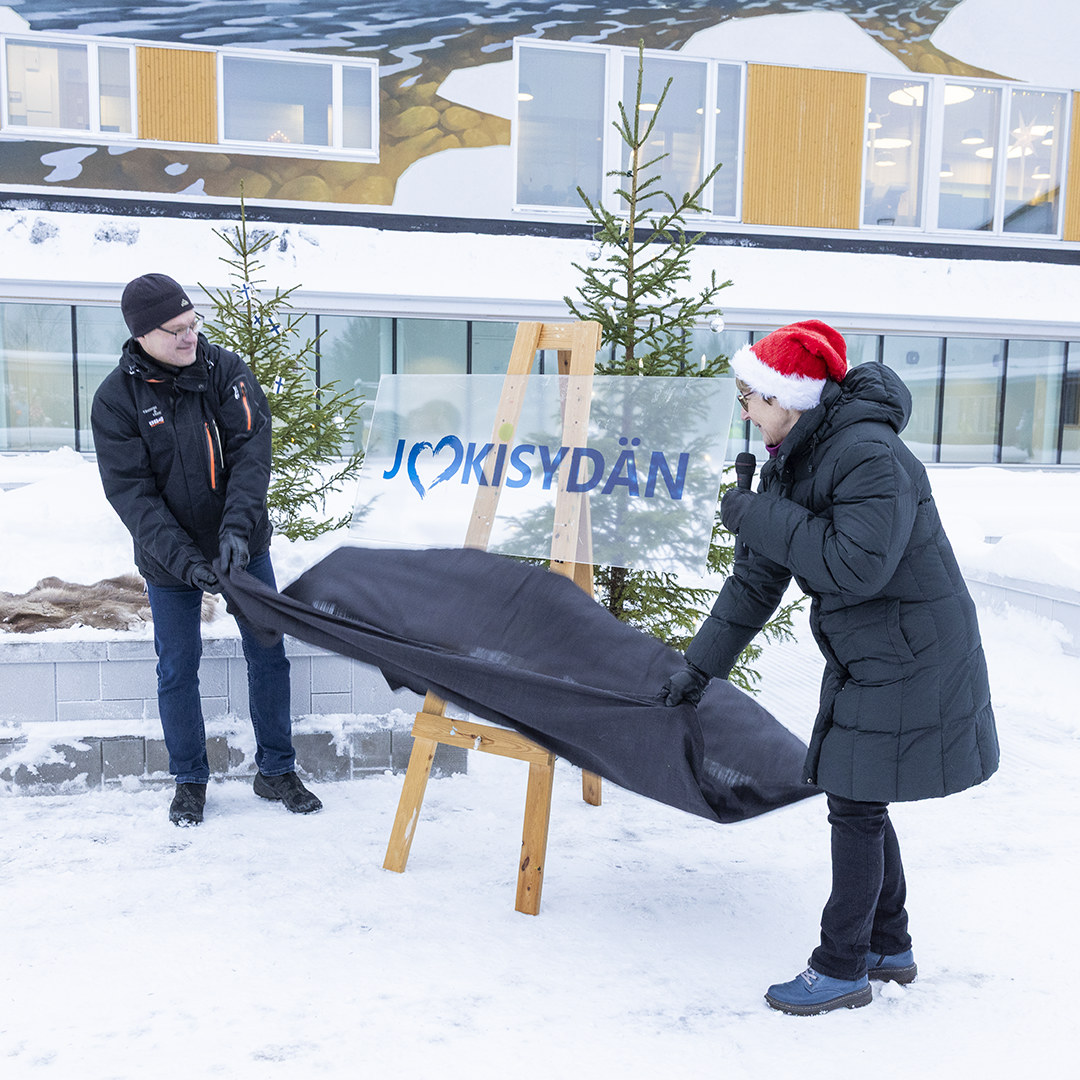 Kaksi henkilöä seisoo lumisessa ulkoilmassa ja nostaa mustaa kangasta paljastaakseen kyltin, jossa lukee ”JOKISYDÄN” sinisellä tekstillä ja sydänsymbolilla. Kyltin alla on puinen teline, ja taustalla näkyy kaksi koristeltua kuusta sekä moderni rakennus, jossa on suuria ikkunoita ja seinämaalauksia. Yksi henkilöistä pitää punaista joululakkia.
