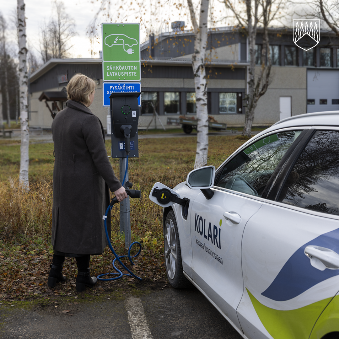 Henkilö lataa sähköautoa latauspisteellä. Auton kyljessä lukee "KOLARI" ja "Kasvaa luonnostaan". Latauspisteessä on vihreä kyltti, jossa on valkoinen sähköauton kuva ja teksti "SÄHKÖAUTON LATAUSPISTE". Sen alla on sininen kyltti, jossa lukee "PYSÄKÖINTI SALLITTU LATAUKSEN AJAKSI". Taustalla näkyy rakennus ja koivupuita.