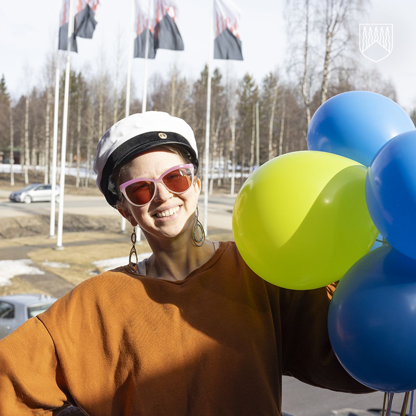 Kuvassa henkilö ylioppilas lakki päässään, ilmapalloja käsissään, aurinkolasit silmillä. Taustalla Kolarin kunnan vaakunaliput, jotka liehuvat lipputangoissa.