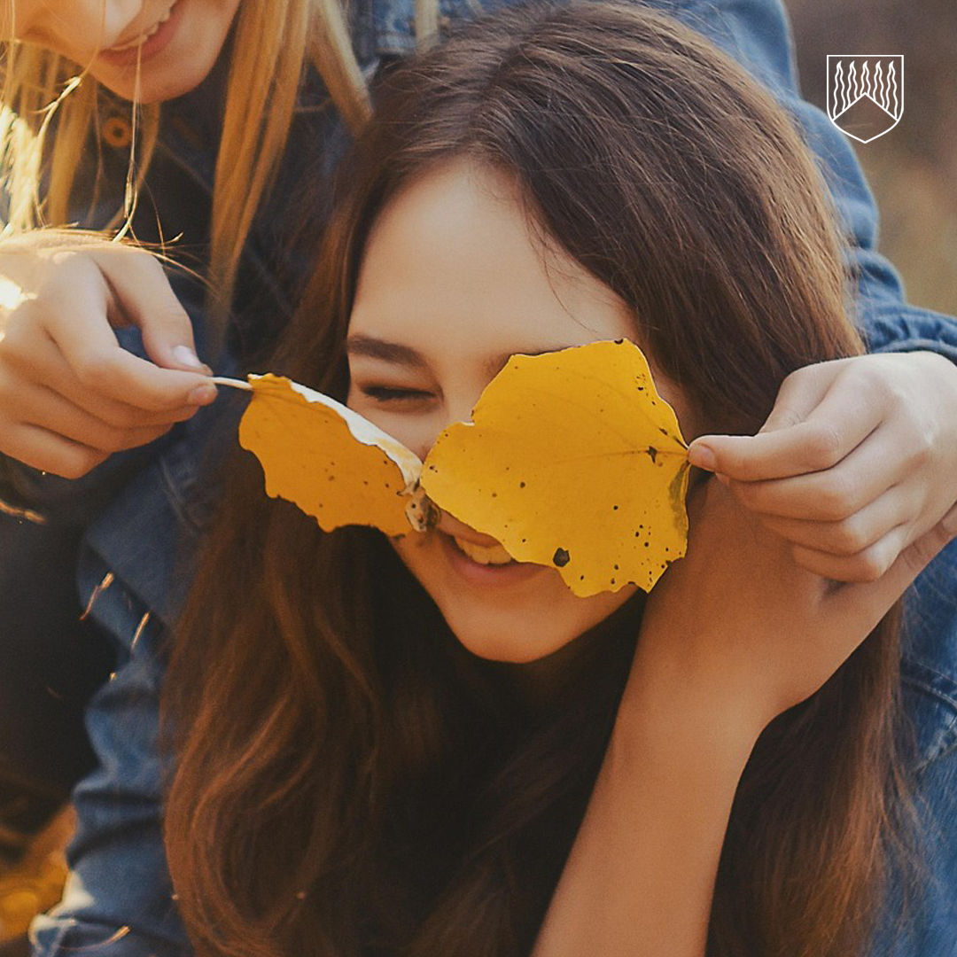 Henkilö pitää kahta keltaista syksyn lehteä toisen henkilön silmien edessä, ikään kuin silmälaseina. Toinen henkilö hymyilee. Tausta on epätarkka.