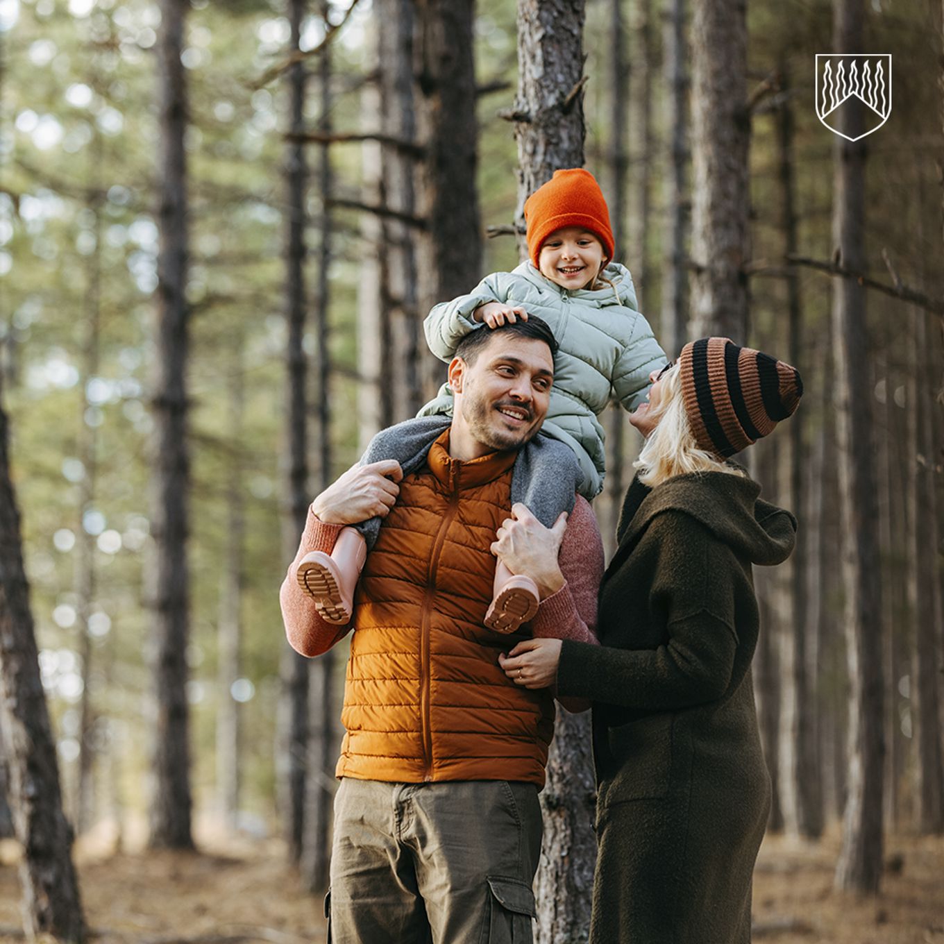 Kolmihenkinen perhe kävelee metsässä. Yksi henkilö kantaa lasta olkapäillään, ja toinen henkilö kulkee vieressä pitäen kantajan kädestä. Metsässä on korkeita, suorarunkoisia puita ja vähän aluskasvillisuutta. Aurinko siivilöityy puiden lomasta, ja kaikilla on päällään lämpimät takit ja pipot.