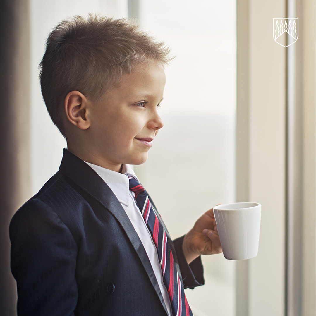 Lapsi pukeutuneena tummansiniseen pukuun, valkoiseen kauluspaitaan ja puna-siniraidalliseen solmioon, seisoo ikkunan edessä ja pitää kädessään valkoista kahvikuppia. Taustalla näkyy vaalea ikkuna.