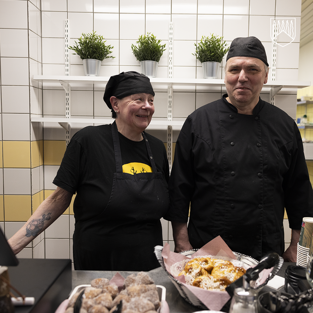 Kaksi kahvilan työntekijää seisoo tiskin takana mustissa työasuissaan. Heidän edessään on esillä leivonnaisia ja muita tarjottavia. Taustalla on valkoisia laattaseiniä ja hyllyillä vihreitä ruukkukasveja.