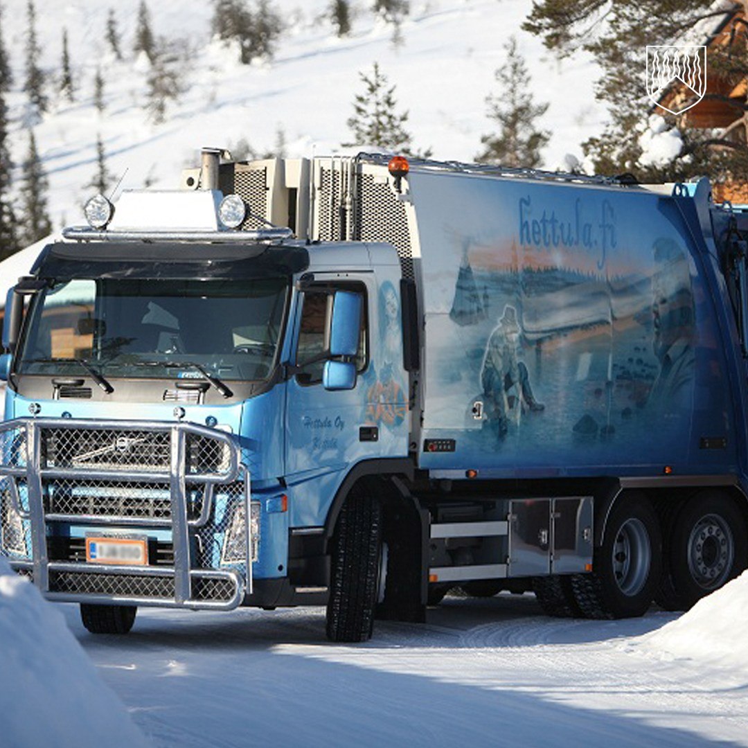 Sinisävyinen jäteauto seisoo lumisella tiellä talvisessa metsämaisemassa. Ajoneuvon kyljessä on taiteellinen maalaus, jossa näkyy jääkarhuja ja arktinen maisema. Taustalla on lumisia puita ja rakennuksen nurkka.