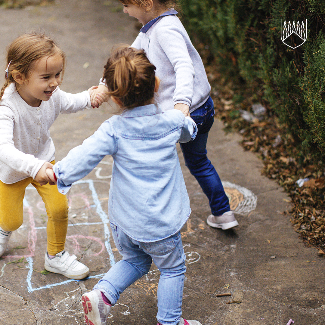 Kolme lasta leikkii pihalla pitäen toisiaan kädestä liidulla piirrettyjen ruutujen päällä.