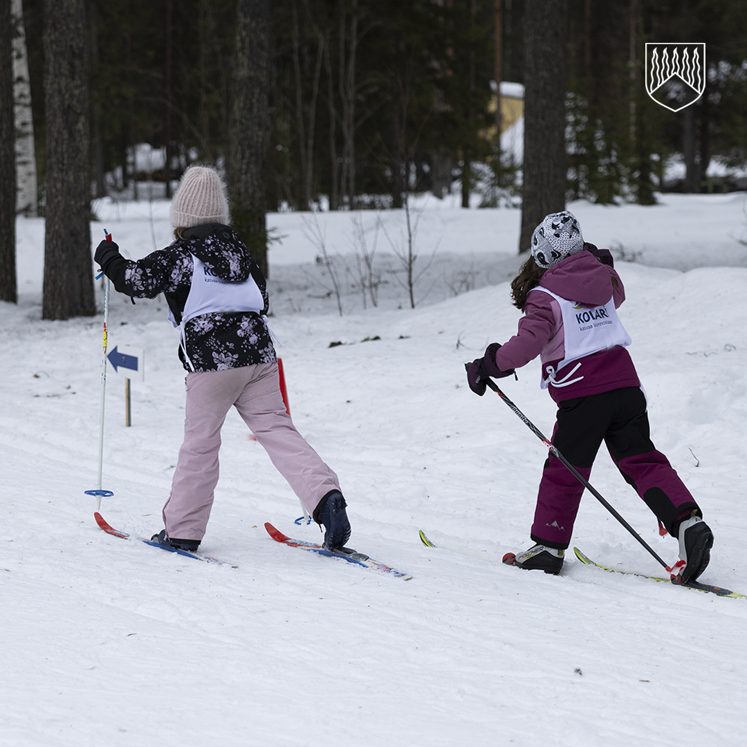 Kaksi hiihtovälineisiin pukeutunutta lasta etenee suksilla lumisella metsäpolulla. Molemmilla on yllään kilpailuliivit ja kuvan taustalla näkyy puita sekä luminen metsämaisema.