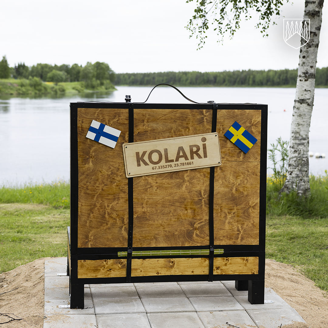 Järven rannalle sijoitettu puinen matkalaukkua muistuttava veistos. Laukun etupuolella on kyltti, jossa lukee ”KOLARI” sekä paikkakunnan koordinaatit. Laukun kannessa on Suomen ja Ruotsin liput, ja taustalla näkyvät järvi, nurmialue ja koivu.