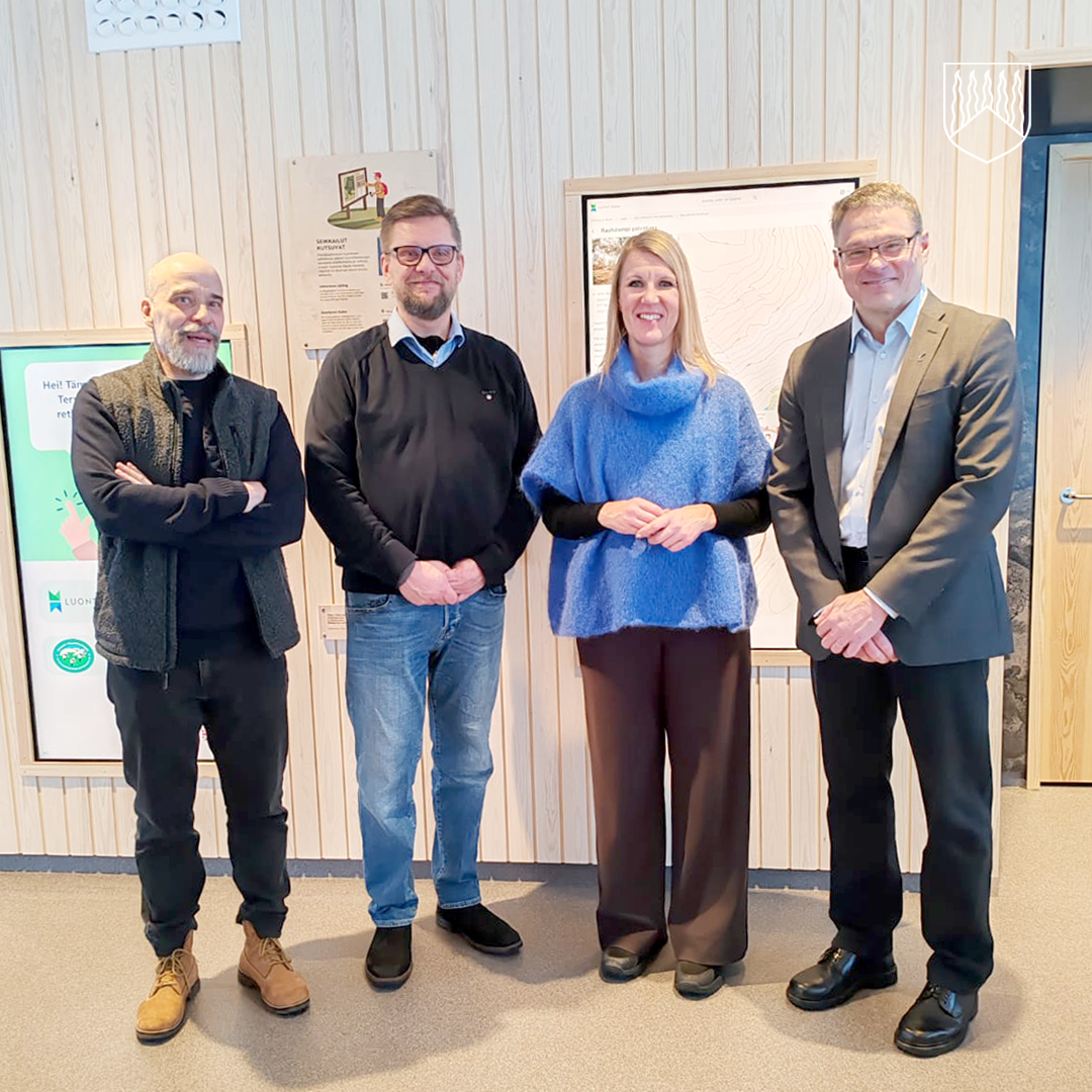 Neljä henkilöä seisoo sisätiloissa vierekkäin, taustallaan vaalea puupaneeliseinä, informaationäyttö ja seinälle kiinnitetty kartta. Henkilöt ovat pukeutuneet siististi ja katsovat kameraan. Tilassa on valoisa ja virallinen tunnelma.