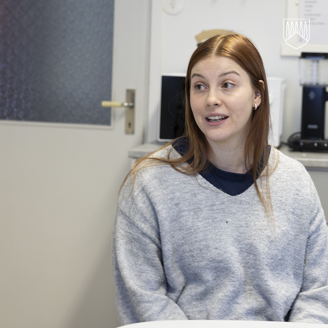 Henkilö istuu sisätilassa pöydän ääressä, taustalla näkyy ovi ja toimistolaitteita.