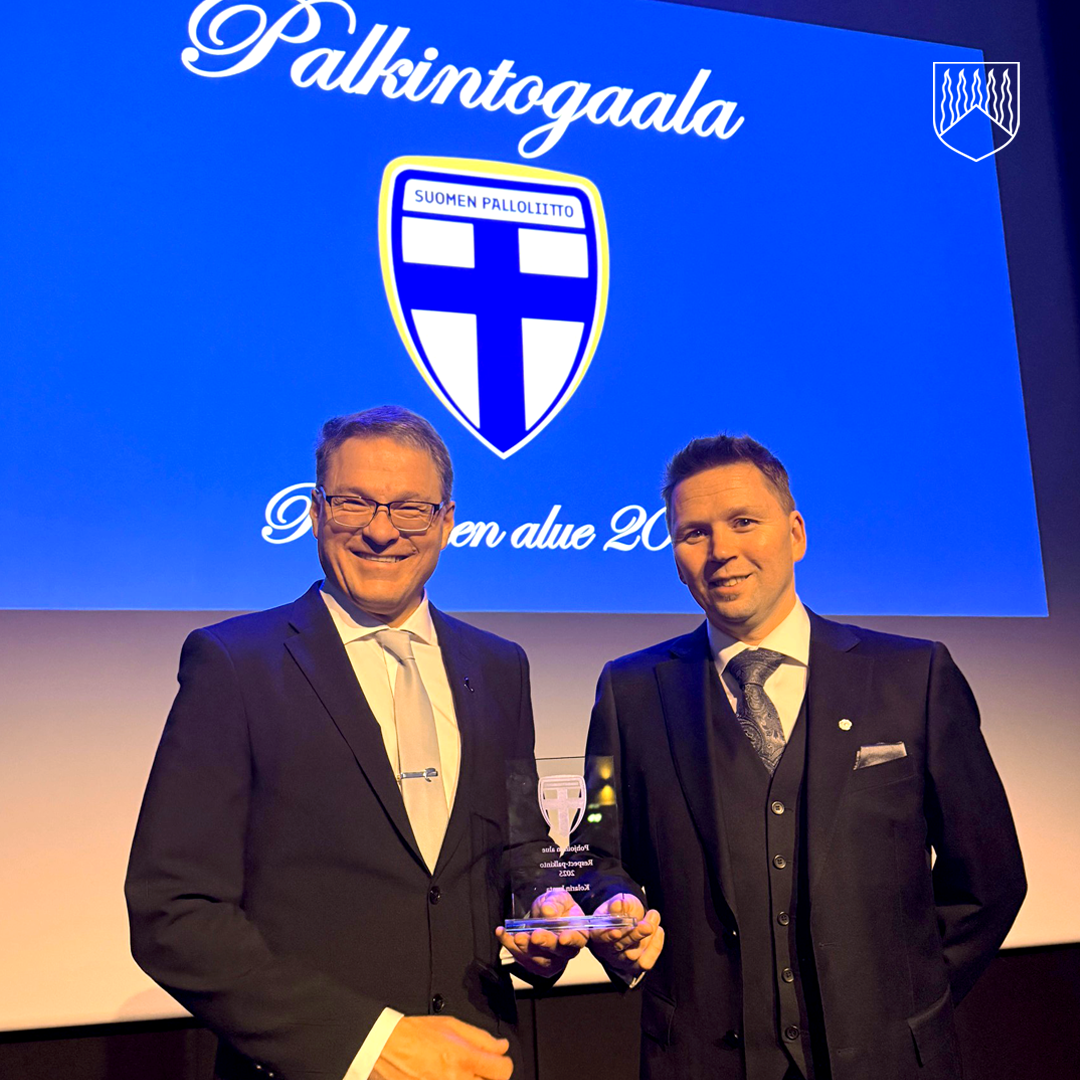 Kaksi pukuihin pukeutunutta henkilöä poseeraa lavalla palkinnon kanssa. Heidän takanaan näkyy suuri sinisävyinen taustakuva, jossa on Suomen Palloliiton logo ja teksti ”Palkintogaala” sekä tapahtuman alueellinen vuosimerkintä. Tilaisuus näyttää olevan virallinen palkinnonjakotapahtuma.
