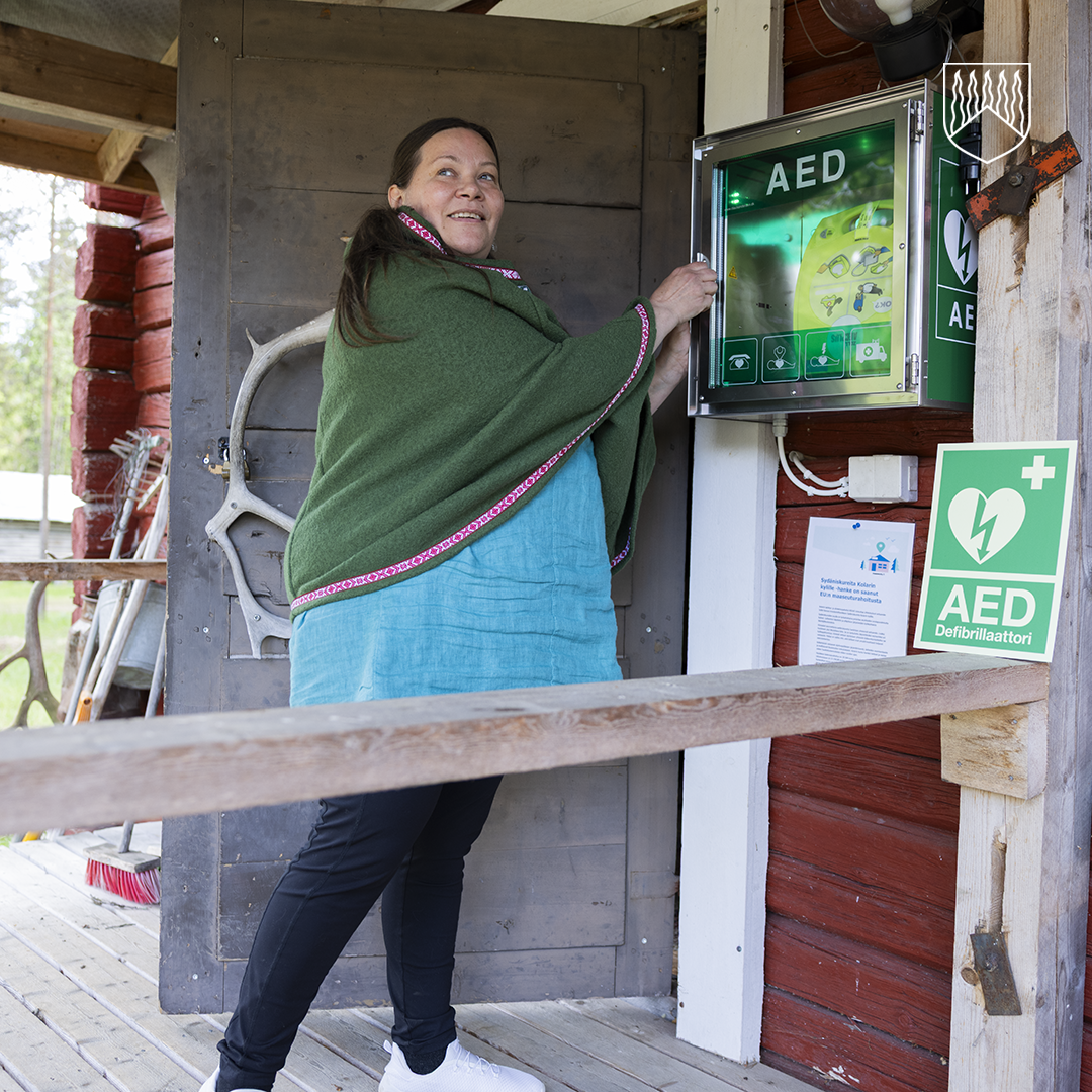 Henkilö avaa rakennuksen seinälle asennettua sydäniskurikaappia. Sydäniskuri on valaistussa AED-kaapissa kuistin seinällä. Kuvassa näkyy myös useita AED-opasteita sekä rakennuksen puinen ulkoseinä ja kuisti.