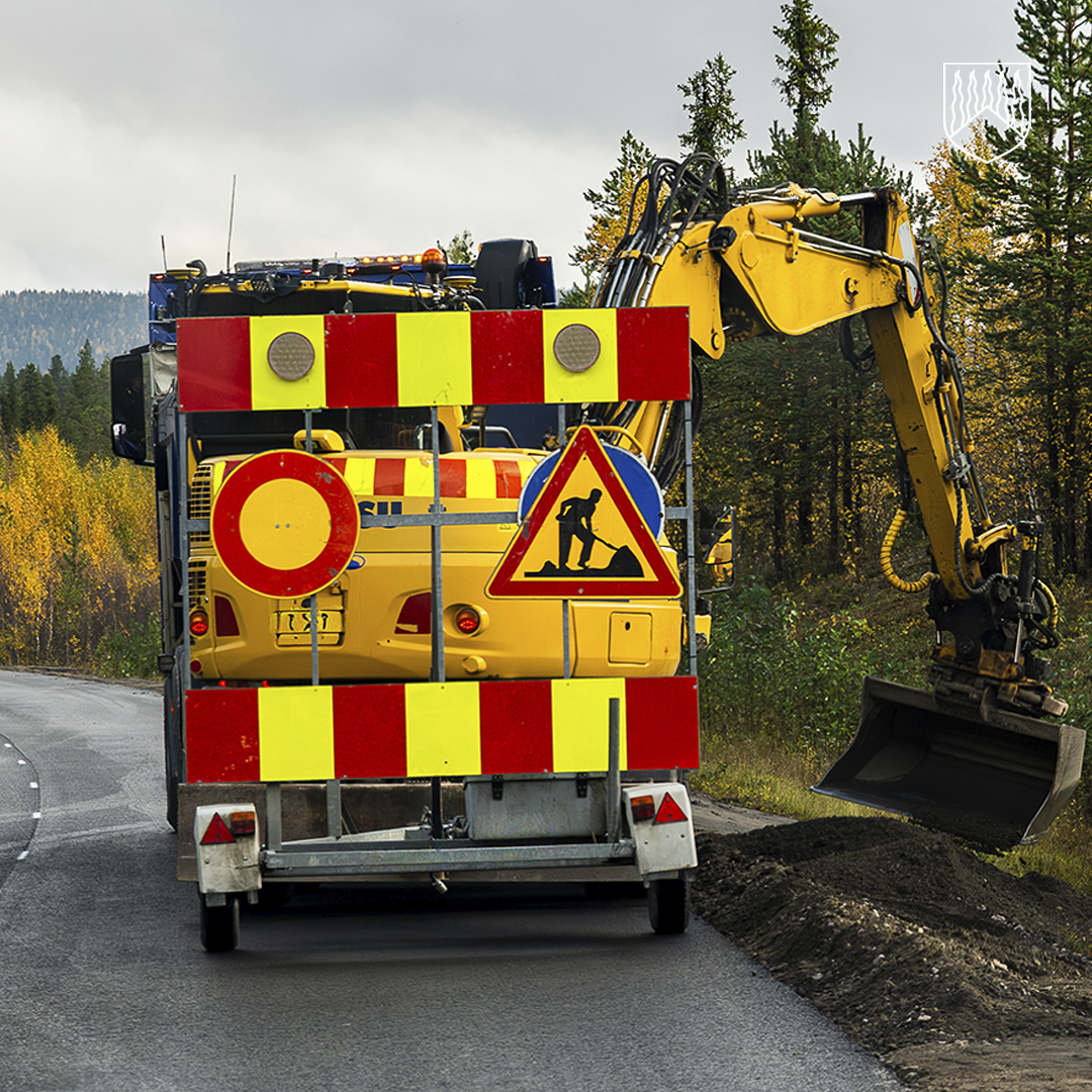 Tietyömaa maantien varressa. Kuvassa näkyy varoitusmerkein varustettu työajoneuvo ja kaivinkone, joka työskentelee tien reunassa metsäisessä ympäristössä.