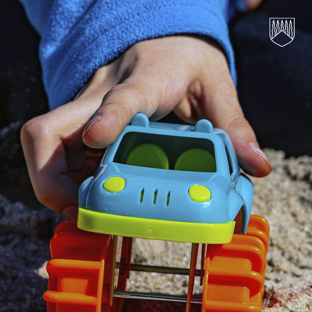 Lähikuva kädestä, joka pitelee sinistä ja vihreää leluautoa, jossa on suuret oranssit pyörät, karkealla kivipinnalla.