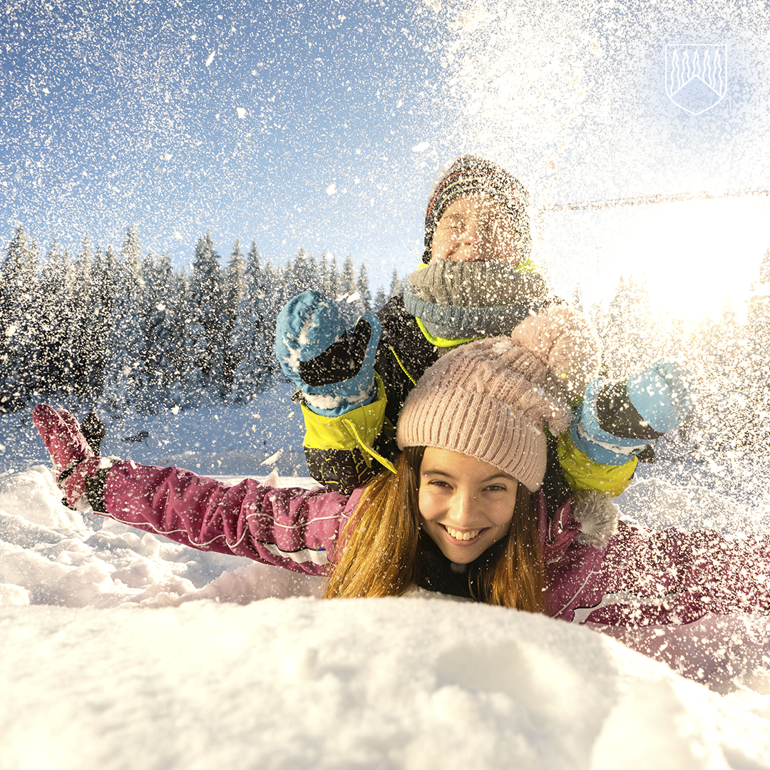 Kaksi lumisiin ulkoiluvaatteisiin pukeutunutta henkilöä leikkii lumessa kirkkaassa auringonpaisteessa. Etualalla oleva henkilö makaa lumihangessa kädet levällään, ja toinen henkilö on heidän takanaan heittämässä lunta ilmaan. Taustalla näkyy lumisia puita ja sininen taivas.