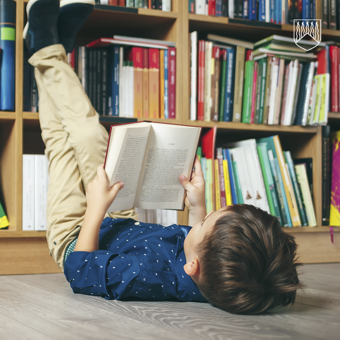 Lattialla makaava lapsi lukee kirjaa jalat nostettuina kirjahyllyä vasten. Taustalla on täysi kirjahylly, jossa on erivärisiä kirjoja.