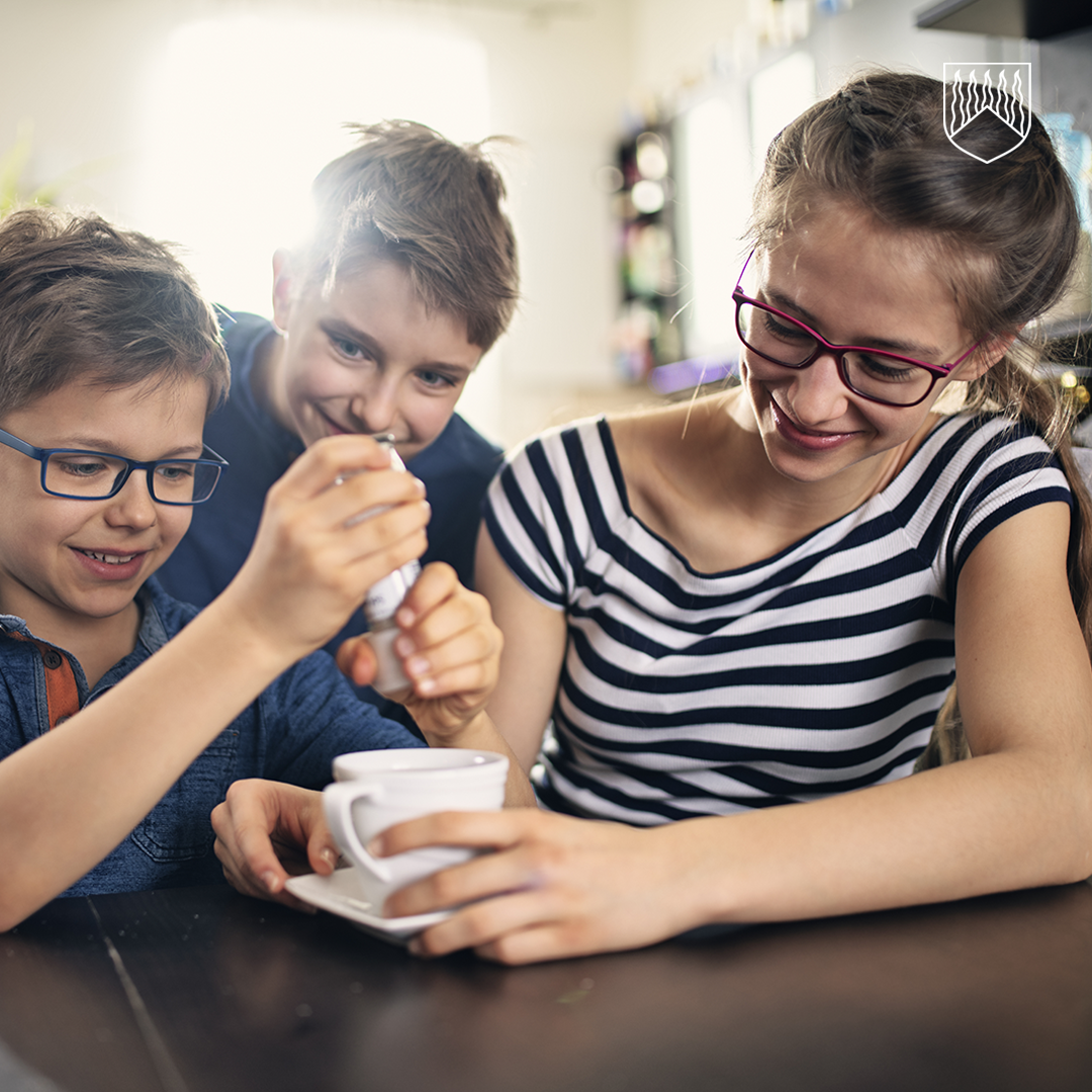 Kolme lasta istuu pöydän ääressä sisätilassa. Lapset työskentelevät yhdessä valkoisen kahvikupin parissa, johon yksi heistä sekoittaa jotakin lusikalla. Kaikilla lapsilla on silmälasit ja he ovat lähellä toisiaan, pöydän äärellä. Tausta on pehmeästi valaistu ja epäselvä.