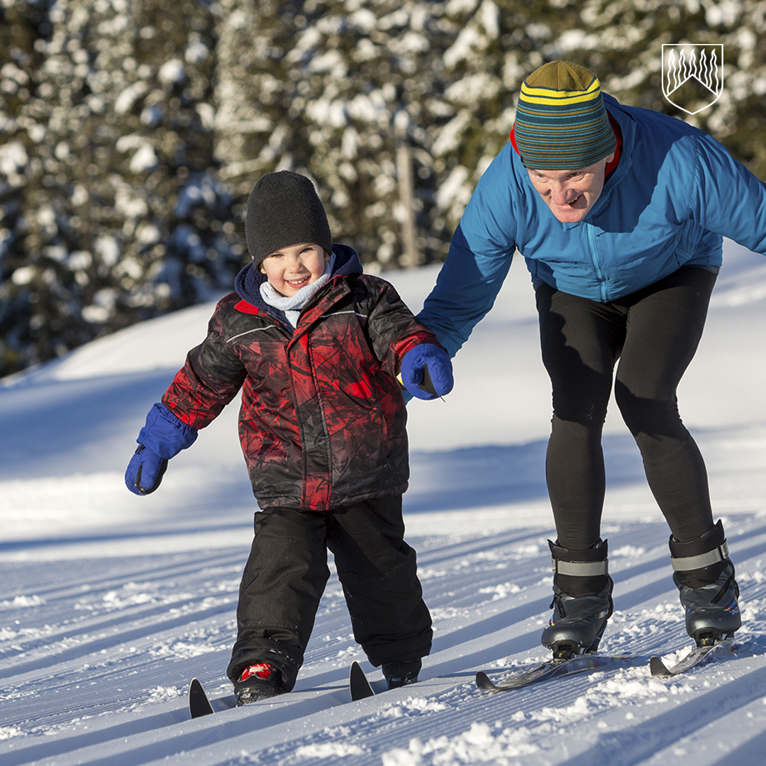 Aikuinen ja lapsi hiihtävät yhdessä aurinkoisella, lumisella metsäpolulla. Aikuinen pitää lasta kädestä ja ohjaa tämän hiihtoa. Molemmilla on talvivaatteet, ja taustalla näkyy lumipeitteisiä puita.