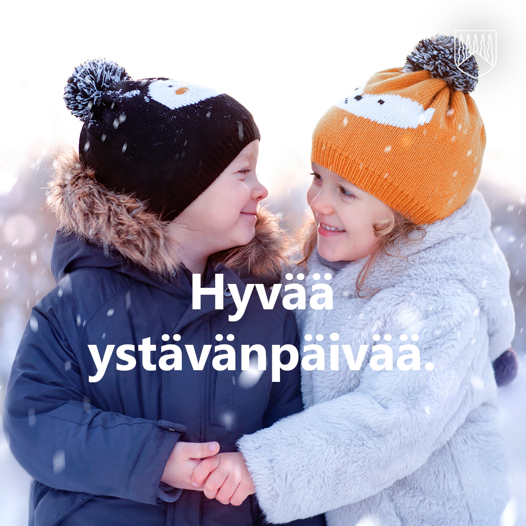 Kaksi talvivaatteisiin pukeutunutta lasta seisoo lumisateessa ja pitävät toisiaan kädestä. Heillä on päässään värikkäät tonttulakkia muistuttavat pipot, ja taustalla näkyy pehmeästi sumeneva talvimaisema. Kuvan keskellä on teksti ”Hyvää ystävänpäivää."