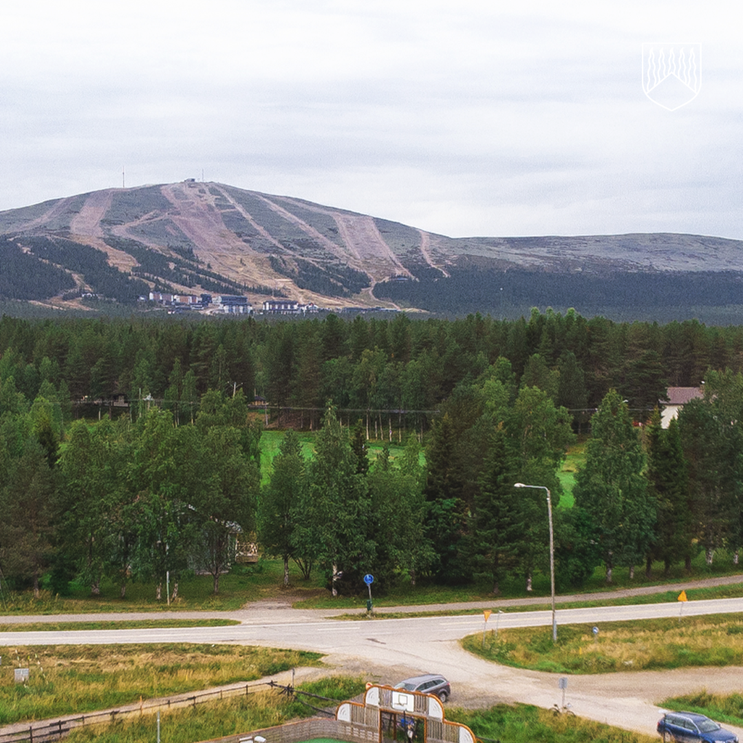 Maisemakuva Ylläksen tunturista kesäaikaan. Etualalla kulkee tie ja risteys, niiden takana näkyy tiheää vihreää metsää. Taustalla kohoaa Ylläksen tunturi, jonka rinteillä erottuvat leveät laskettelurinteet ja niiden juurella matkailukeskuksen rakennuksia. Taivas on pilvinen ja valo pehmeä.